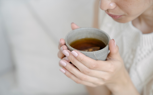 Woman holding a ceramic cup of tea near her face, showing pink nails and a soft neutral indoor background