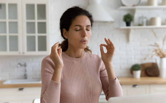 Woman practicing calming breathwork with eyes closed and hands in a mudra in a bright kitchen