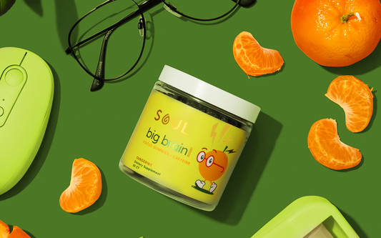 Angled jar of Soul Big Brain tangerine gummies with label and white lid, tangerine slices and glasses on green background