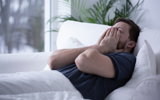 Man lying on a white couch covering his face with both hands, appearing stressed in a bright living room with houseplants