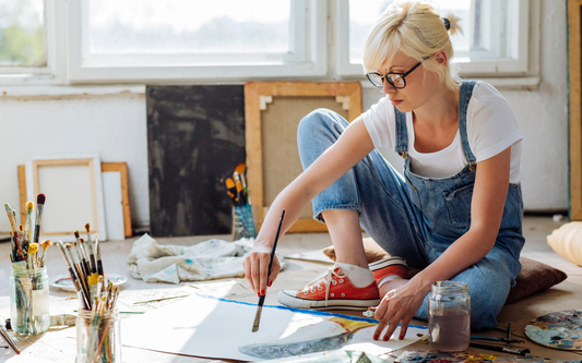 Young woman painting on paper on the floor of a sunlit art studio, surrounded by jars of brushes and stacked canvases