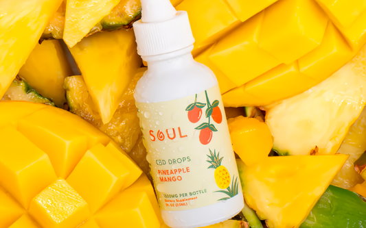 Angled close-up of Soul CBD Drops bottle showcasing Pineapple Mango flavor label and water droplets with diced mango and pineapple background