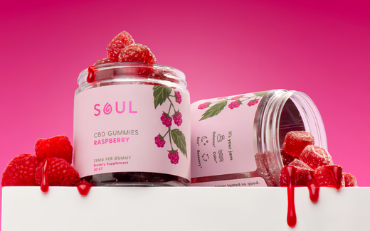 Front view of Soul Raspberry CBD gummy jars, one upright and one tipped, showing label and sugar-coated gummies on a bright pink background
