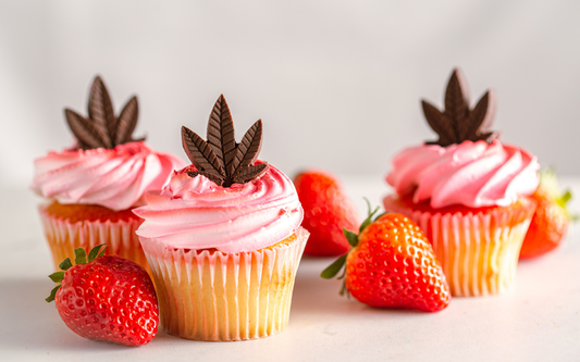 Three cupcakes with pink frosting topped with chocolate cannabis-leaf decorations and fresh strawberries on a light background