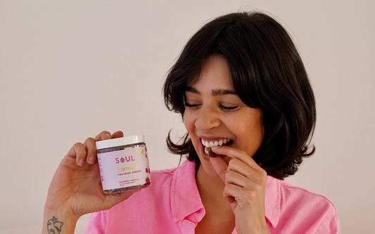 Woman smiling while eating a gummy and holding a jar of Soul Harmoni PMS Relief Gummies against a neutral background