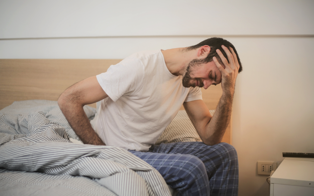 Hombre sentado en la cama sosteniéndose la cabeza y la cintura con expresión de dolor en un dormitorio