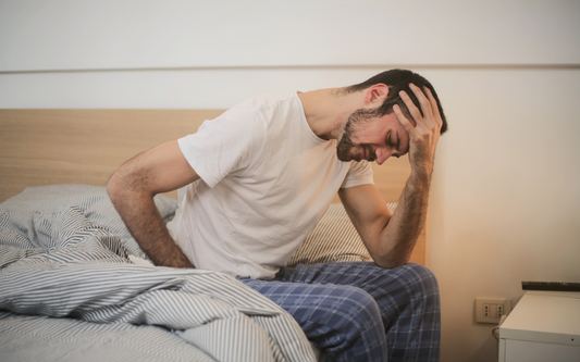 Hombre sentado en la cama sosteniéndose la cabeza y la cintura con expresión de dolor en un dormitorio