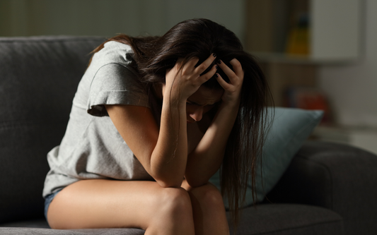 Mujer joven sentada en un sofá con la cabeza entre las manos, mostrando ansiedad en un entorno doméstico con luz tenue