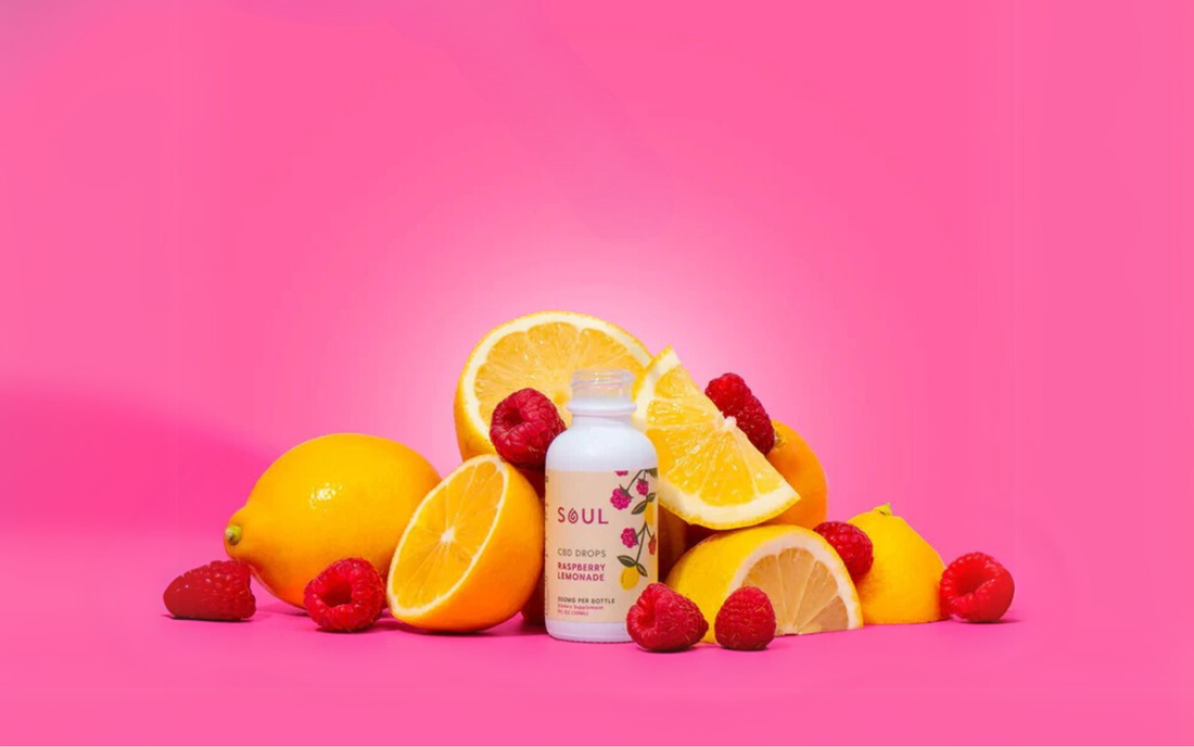 Vista frontal de frasco Soul CBD drops con etiqueta Raspberry Lemonade, rodeado de limones y frambuesas sobre fondo rosa