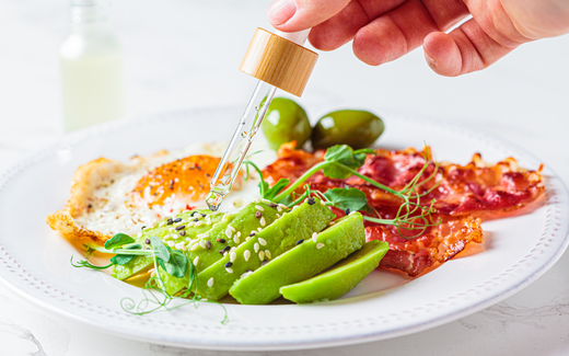 Hand using a dropper to add oil to a plate with sliced avocado, fried egg, crispy bacon, and olives on a white marble surface