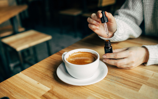 Hands holding a CBD oil dropper above a cup of coffee on a wooden café table