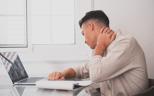 Man at a desk holding his neck in pain beside a laptop in a bright home office