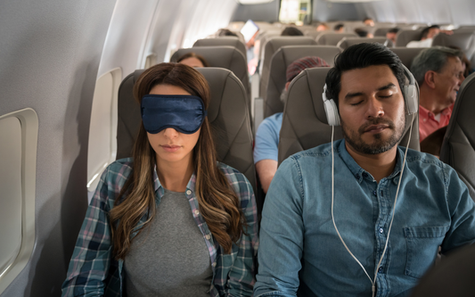 Two airplane passengers sleeping, woman wearing a navy sleep mask and man with headphones in an economy cabin