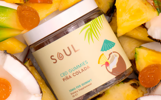 Angled jar of Soul Piña Colada CBD gummies showing 25 mg per gummy label, surrounded by pineapple, coconut, and gummy pieces