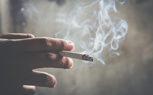 Hand holding a lit joint with curling smoke against a neutral gray background