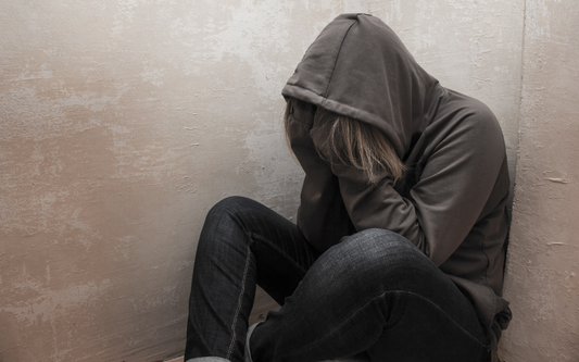 Hooded person sitting on the floor with head in hands, appearing distressed against a textured beige wall