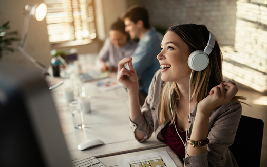 Young woman smiling and listening to music with headphones at a bright shared workspace, colleagues blurred in the background