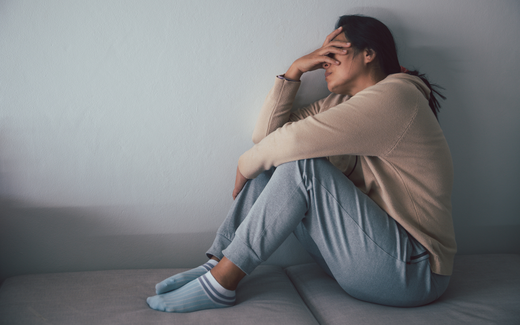 Woman sitting with knees drawn, hand covering her face, appearing distressed against a pale wall