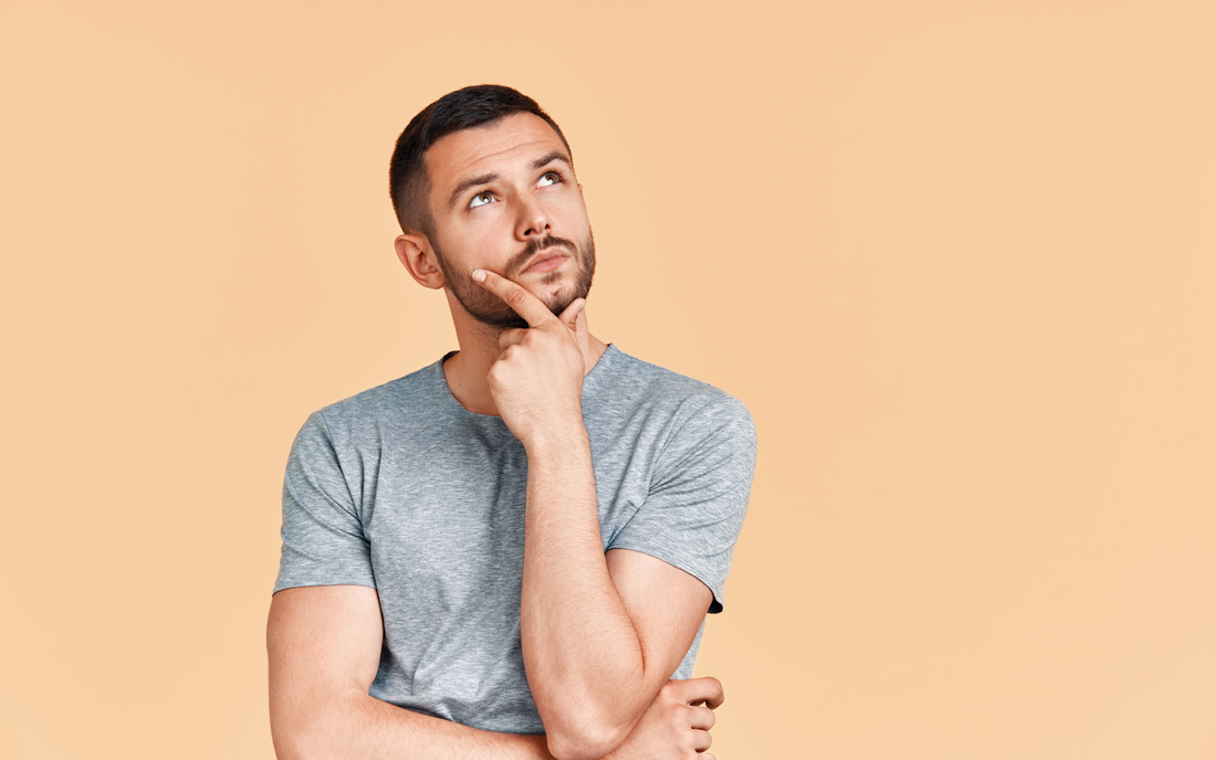 Young man with short dark hair and beard looking thoughtful with hand on chin against a peach background