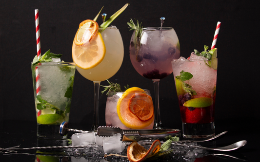 Assortment of five colorful cocktails on a black tabletop, garnished with citrus slices, fresh herbs, and crushed ice