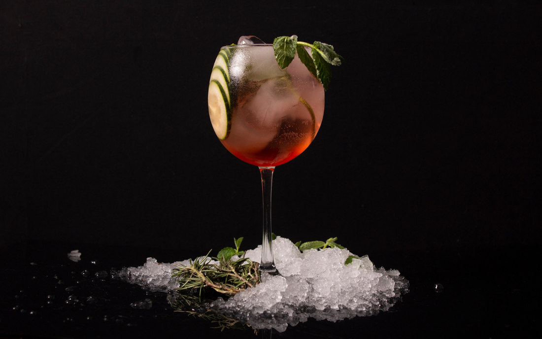 Wine glass with a pink cocktail garnished with cucumber slices and mint, sitting on crushed ice against a black background