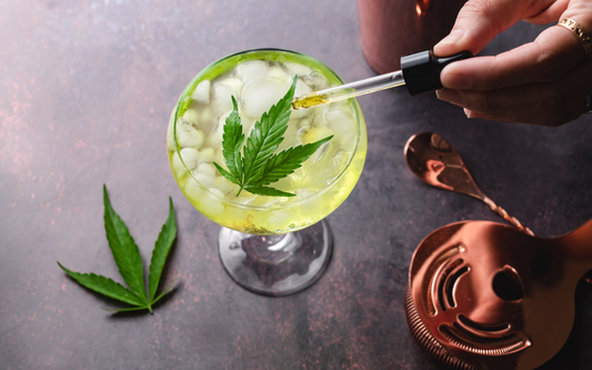 Cocktail glass of iced green drink topped with a cannabis leaf while a dropper adds golden THC oil over it on a dark surface