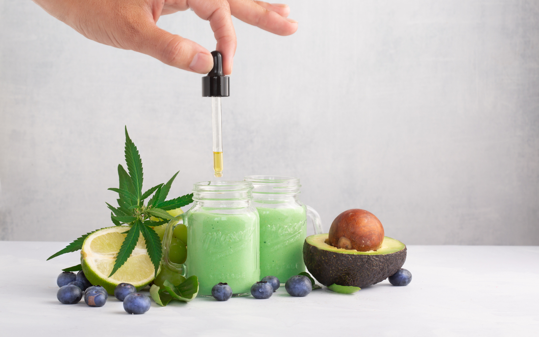 Hand dropping cannabis oil into two green avocado-lime smoothies, surrounded by a cannabis leaf, halved avocado, lime, and blueberries
