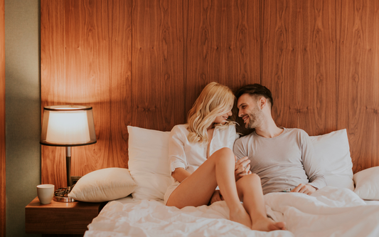 Couple sitting on a bed, holding hands and smiling in a warmly lit bedroom with a wood-paneled headboard