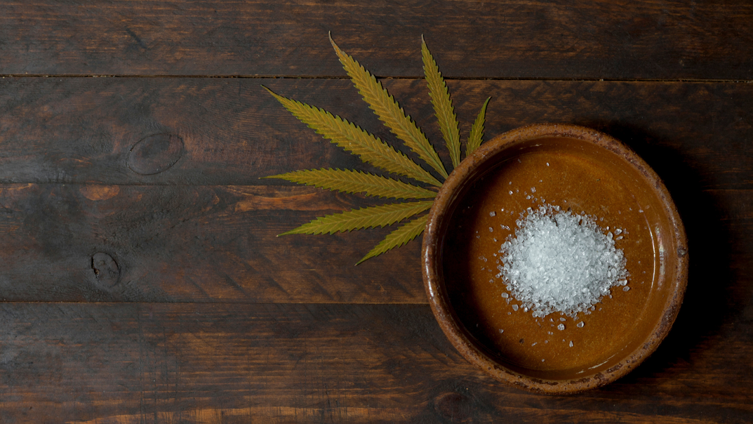 Bowl of white CBD isolate crystals beside a cannabis leaf on a dark wooden table