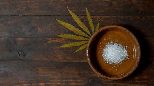 Bowl of white CBD isolate crystals beside a cannabis leaf on a dark wooden table