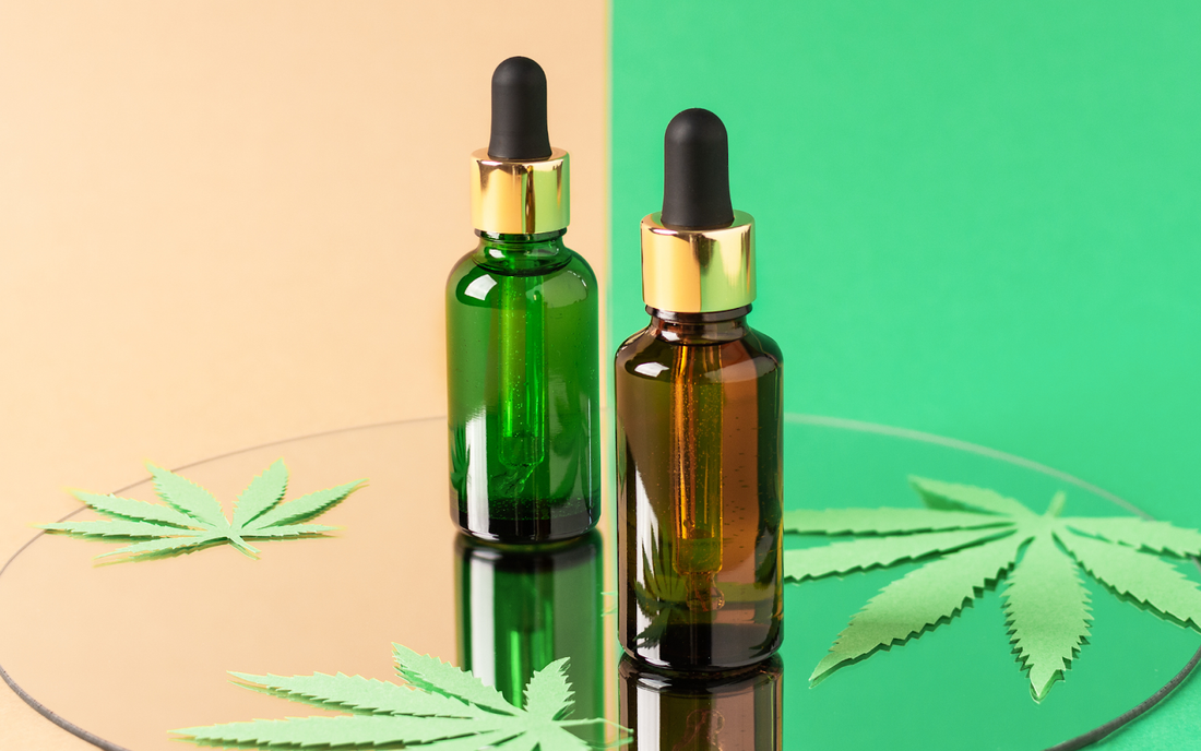 Two CBD oil dropper bottles on a mirror with paper cannabis leaves and split green and beige background