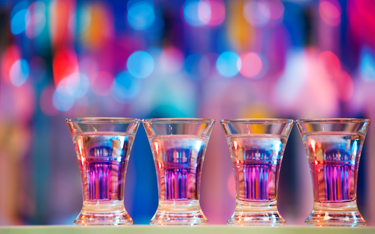 Four shot glasses with purple-hued drinks lined on a bar ledge against colorful blurred bokeh lights