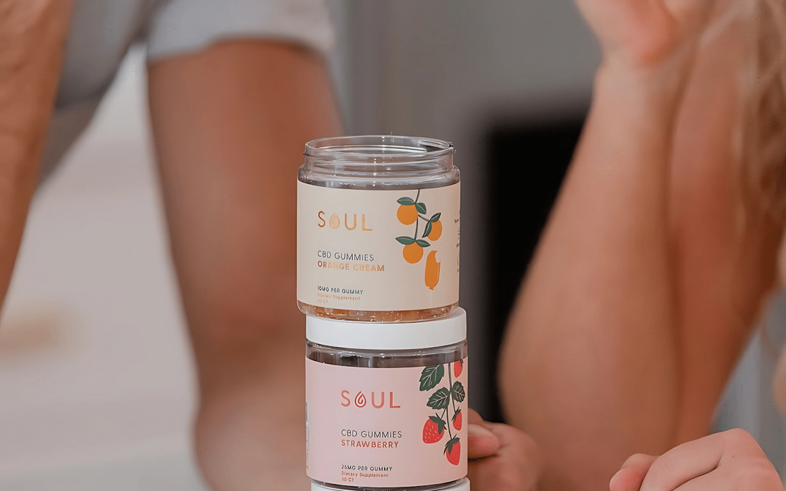 Front view of two stacked Soul CBD Gummies jars — top jar (Orange Cream) with open lid, bottom jar (Strawberry) labeled, on a blurred indoor background