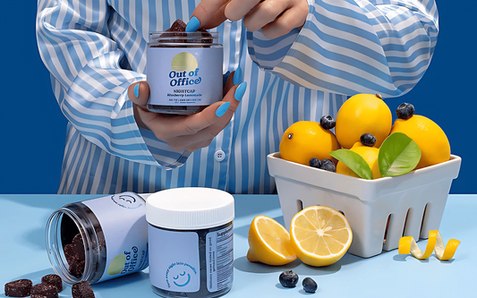 Person in blue striped shirt holding a jar labeled Out of Office gummies over a table with jars, sliced lemon, and blueberries