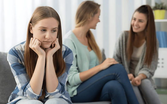 Young woman sitting apart and looking anxious while two women chat and smile in the background