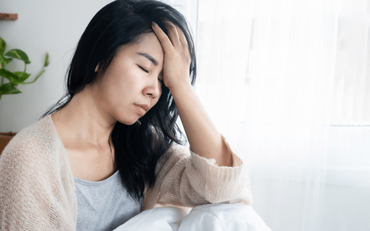 Woman holding her forehead in pain on a bed by a bright window, appearing to have a hangover