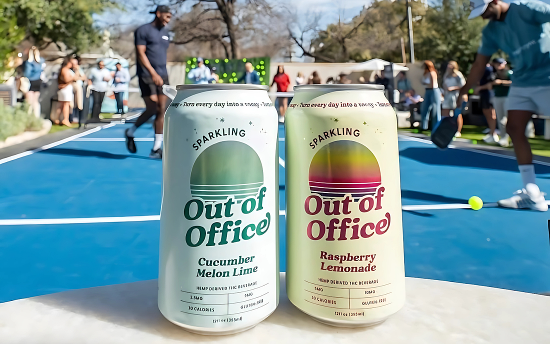 Front view of two Out of Office sparkling cans, Cucumber Melon Lime and Raspberry Lemonade, on a table with blurred pickleball players in the background