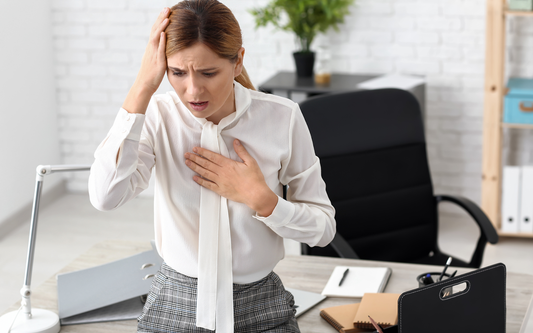 Woman in a white blouse clutching her chest and holding her head at an office desk, appearing anxious