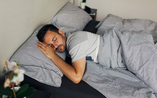 Man sleeping on his side under gray-and-white striped sheets in a calm bedroom setting