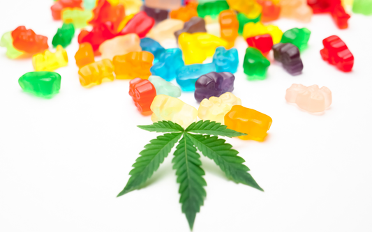 Cannabis leaf resting in front of scattered colorful gummy bear edibles on a white background