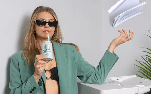 Woman in sunglasses sipping a can labeled Out of Office through a straw while tossing papers above an office copier
