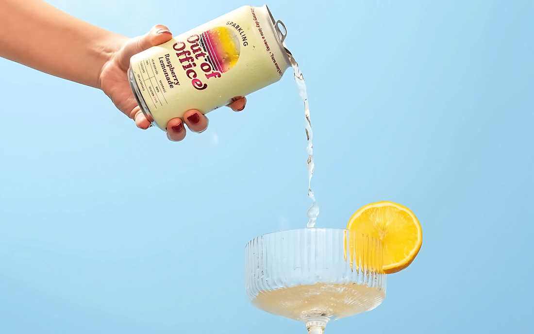 Hand pouring a can of sparkling raspberry lemonade into a coupe glass garnished with a lemon slice against a pale blue background
