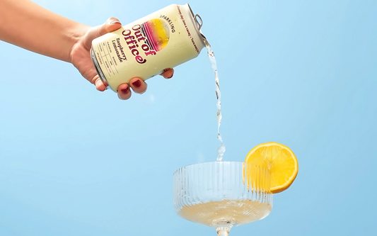 Hand pouring a can of sparkling raspberry lemonade into a coupe glass garnished with a lemon slice against a pale blue background