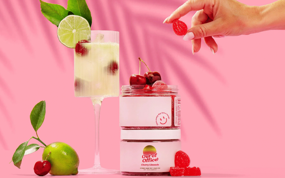 Front view of stacked Out of Office Cherry Limeade gummy jars, one open showing sugar-coated cherry gummies, cocktail glass with lime and cherries on a pink background