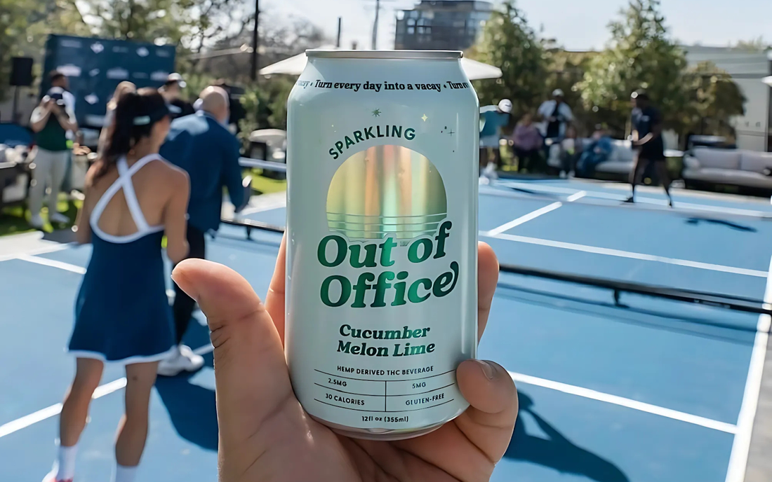 Front view of Out of Office can, Cucumber Melon Lime, held in hand with label visible against blurred pickleball court
