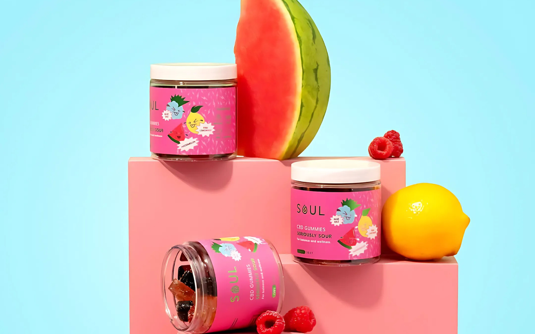 Front view of three Soul CBD gummy jars with pink labels; one tipped open showing gummies, with watermelon, lemon, and raspberries on pink blocks