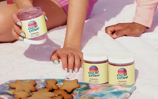 Two people lounging on a white picnic blanket with jars of Out of Office raspberry spread and cookies