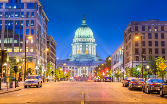 Wisconsin State Capitol building lit at dusk centered at the end of a city street with parked cars and streetlights