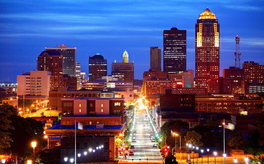 Iowa downtown skyline at dusk with illuminated high-rise buildings and a long, lamp-lined avenue leading into the city