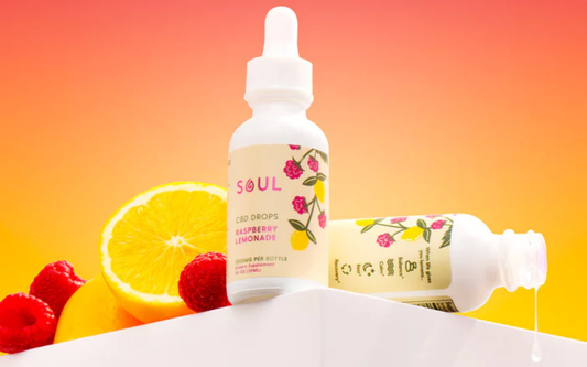 Front of Soul Raspberry Lemonade CBD drops bottle; second bottle pouring oil beside raspberry and lemon on warm gradient